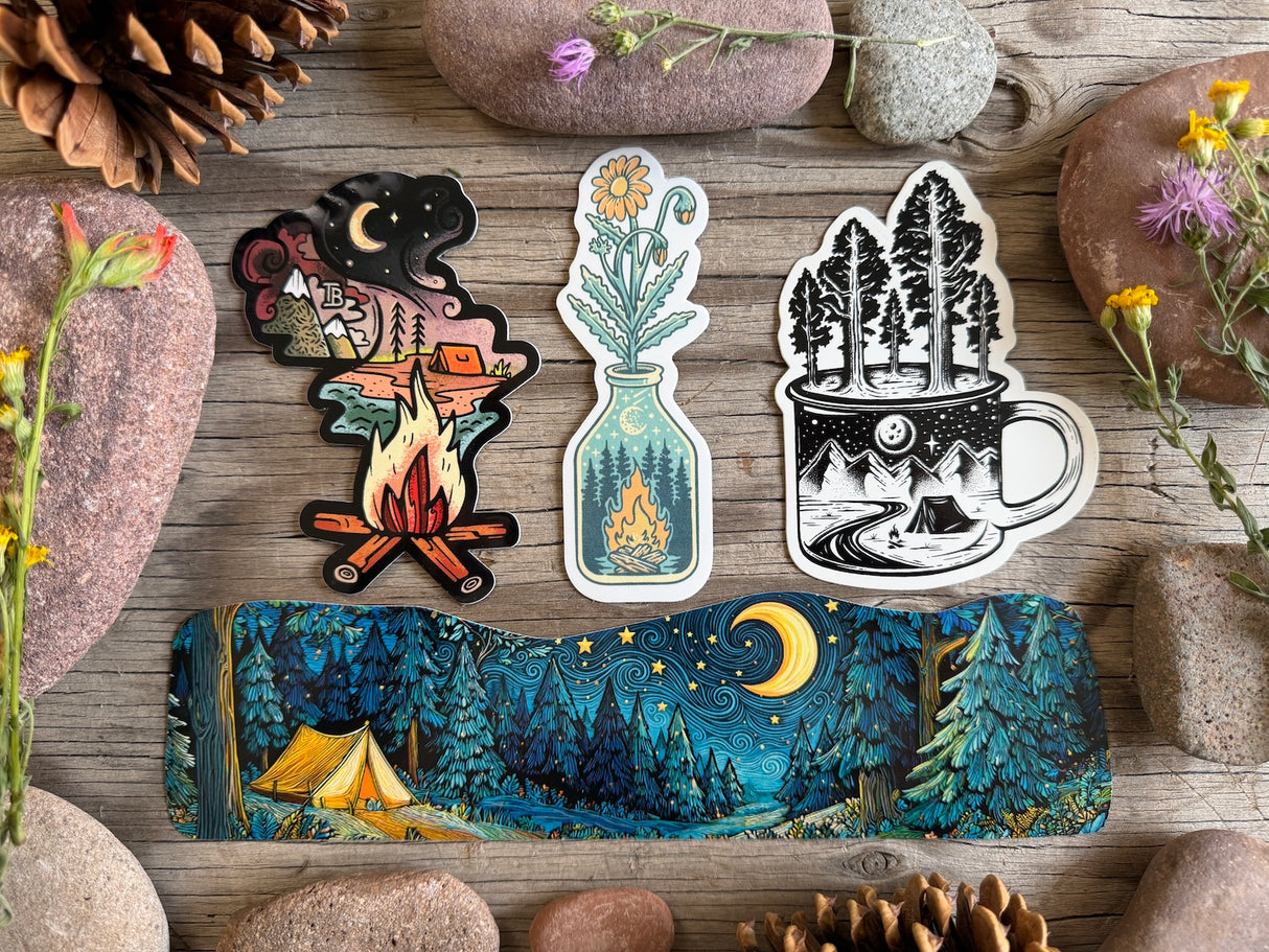 Three die-cut stickers and a long rectangular sticker featuring a camping night scene, arranged on a rustic wooden surface with stones and flowers around them.