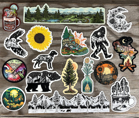Best-selling outdoor stickers by Sticker Art arranged on a wooden surface — silkscreen printed nature designs including mountains, forests, and wildlife