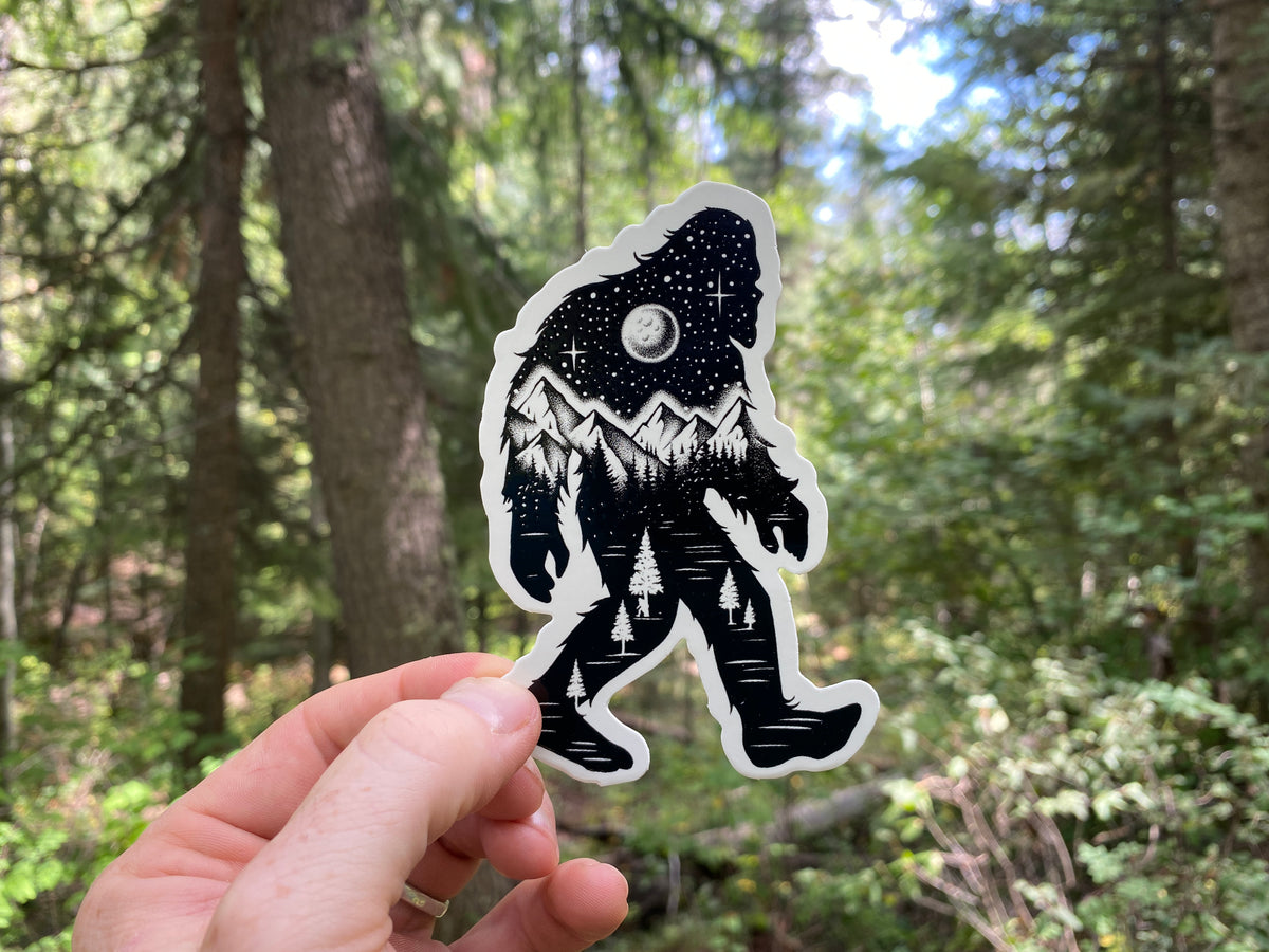 Black-and-white Bigfoot sticker held outdoors in a forest