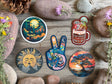 A set of colorful illustrated stickers arranged on a rustic wooden surface with rocks and small flowers. The stickers feature whimsical designs including a moonlit forest, a coffee cup with the words 'FAST COFFEE AND ADVENTURE,' a sun face, a peace sign filled with travel scenes, and a sunset landscape.