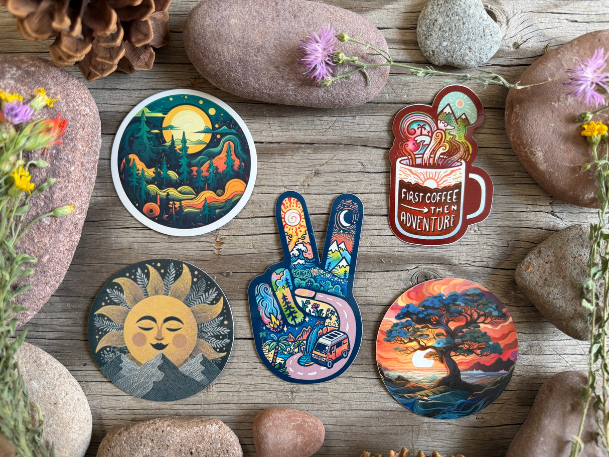 A set of colorful illustrated stickers arranged on a rustic wooden surface with rocks and small flowers. The stickers feature whimsical designs including a moonlit forest, a coffee cup with the words 'FAST COFFEE AND ADVENTURE,' a sun face, a peace sign filled with travel scenes, and a sunset landscape.