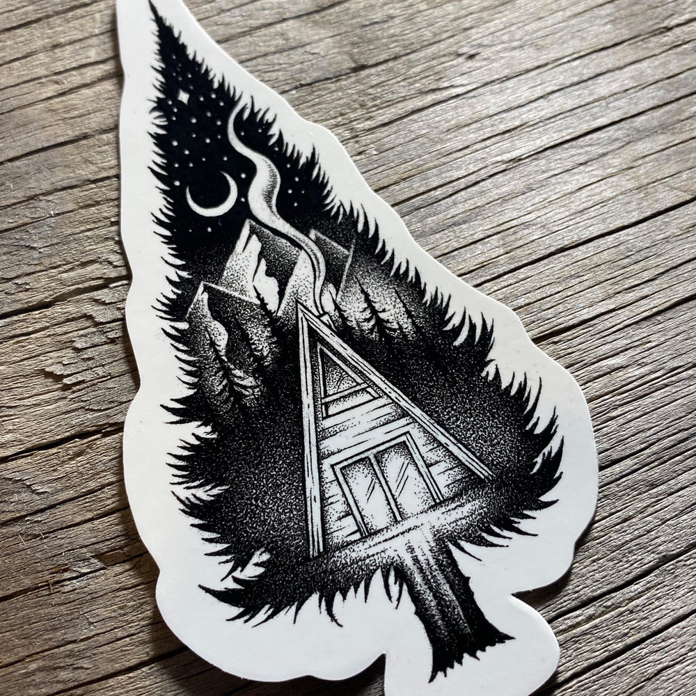 Black-and-white A-frame cabin beneath a crescent moon on wood surface
