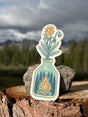 Hand holding the Campfire & Flowers sticker, a wildflower in a glass bottle with a campfire and stars