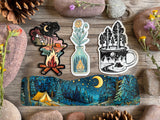 Three die-cut stickers and a long rectangular sticker featuring a camping night scene, arranged on a rustic wooden surface with stones and flowers around them.