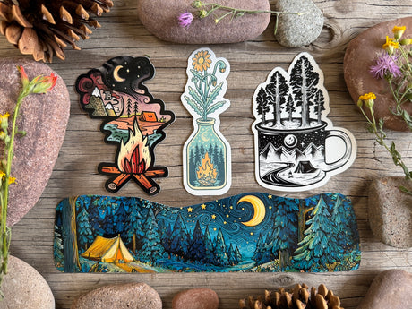 Three die-cut stickers and a long wrap-around sticker featuring a camping night scene, arranged on a rustic wooden surface with stones and flowers around them.