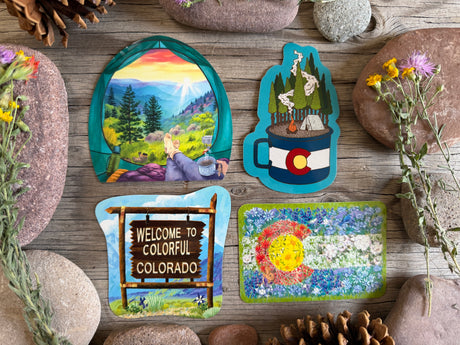 Four colorful Colorado-themed decorative items arranged on a rustic wooden surface with stones and dried flowers. The set includes an arched landscape print showing a person by a lake, a mug-shaped design with a mountain and Colorado flag, a 'Welcome to Colorful Colorado' sign, and a floral patchwork-style square with a sun and blue flowers.