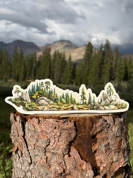 A die-cut wrap-around sticker depicting a mountain and forest landscape, displayed on top of a tree stump with a blurred outdoor mountain background.