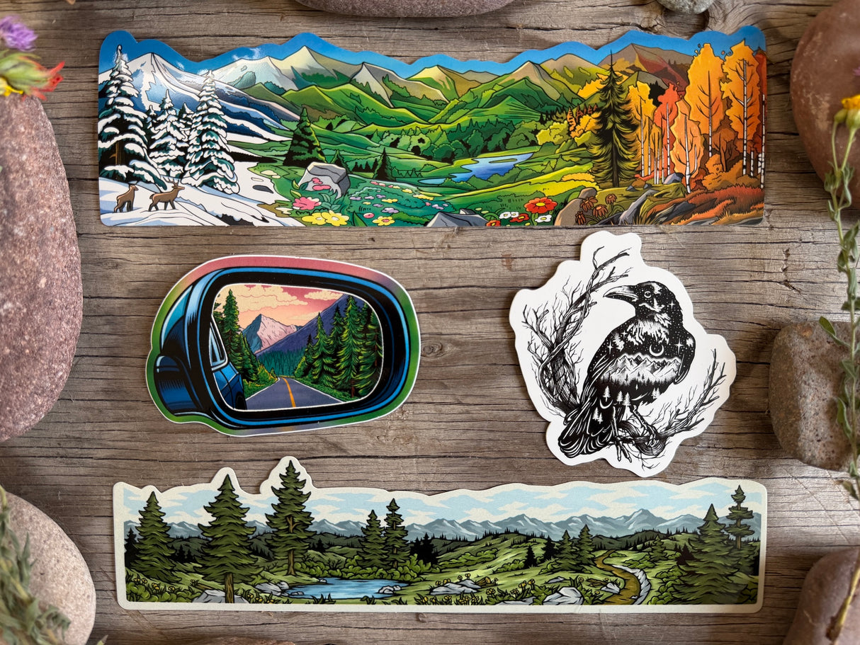 Three die-cut stickers featuring mountain and forest landscapes, arranged on a rustic wooden surface with stones around them. One sticker shows a scenic valley with snowy peaks, evergreen trees, and colorful autumn foliage.