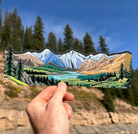 Hand holding a Colorado 14ers water bottle wrap sticker in front of snowcapped Rocky Mountain peaks and pine forest