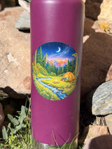 Camping sticker on a water bottle beside river rocks