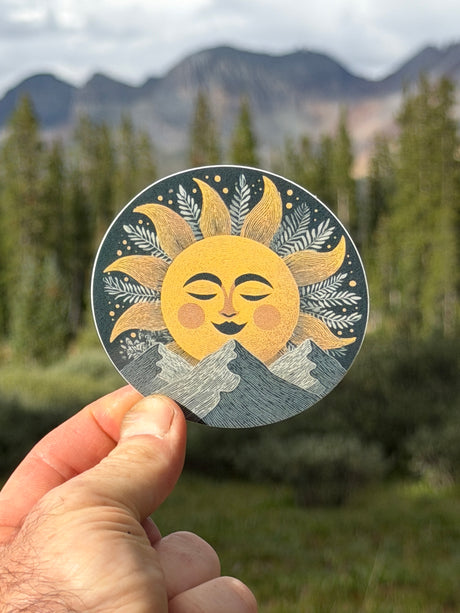 A hand holds a round sticker featuring a smiling sun face with closed eyes, yellow rays, and gray mountains, set against a blurred outdoor background of pine trees and mountains.