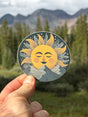 A hand holds a round sticker featuring a smiling sun face with closed eyes, yellow rays, and gray mountains, set against a blurred outdoor background of pine trees and mountains.